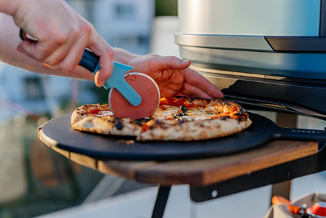 Limited edition pizza cutter featuring a blue handle and red cutting wheel slicing a freshly baked pizza outdoors