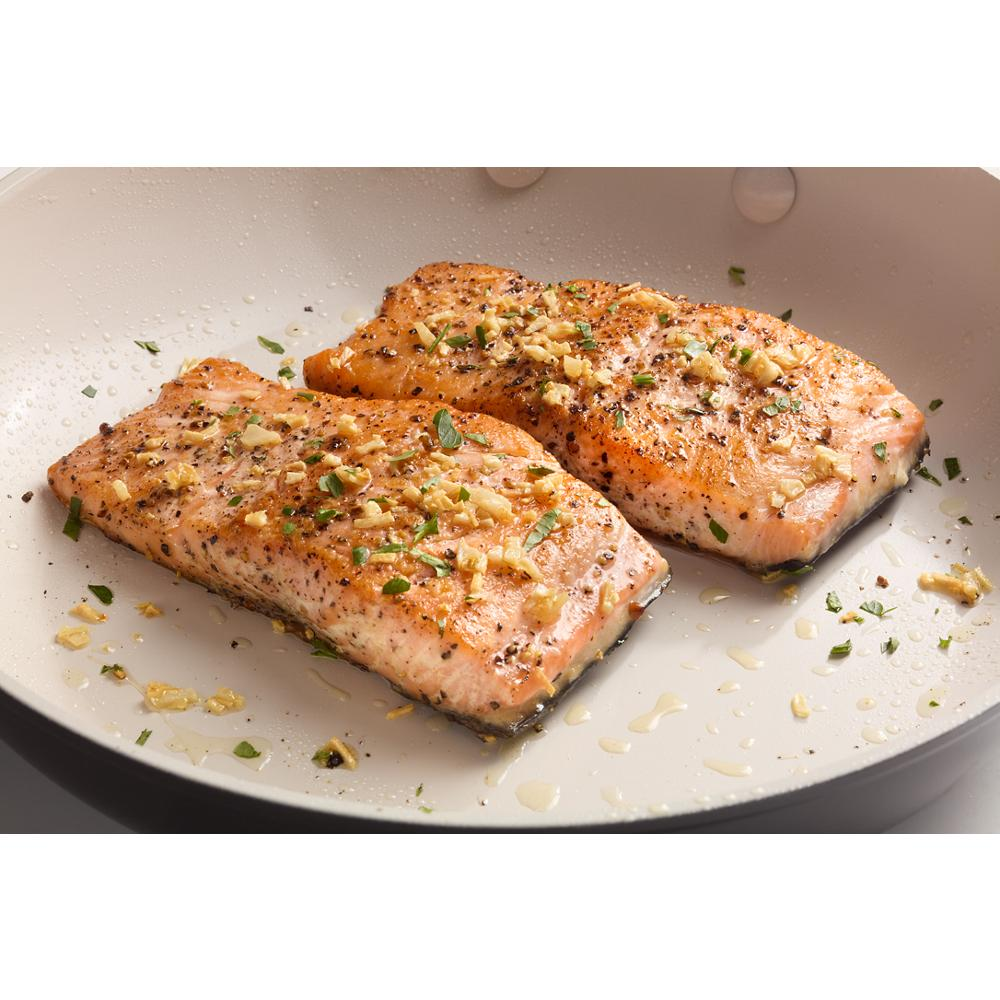 Two seared salmon fillets with herbs and garlic cooking in a light-colored nonstick frying pan