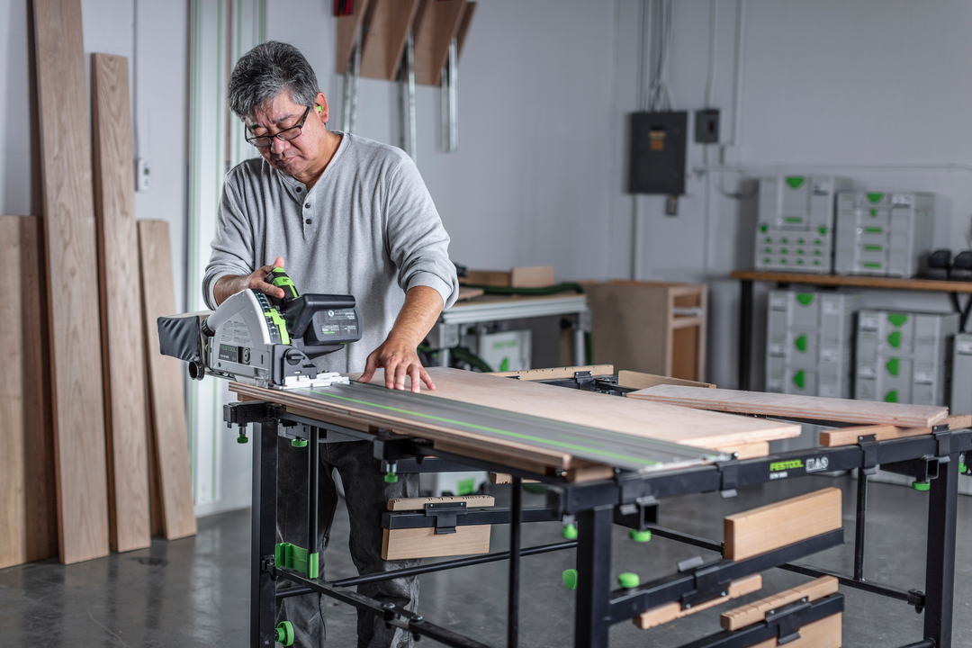 Person using cordless Festool track saw with guide rail cutting wood on workbench in power tools workshop setting