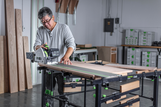 Person using cordless Festool track saw with guide rail cutting wood on workbench in power tools workshop setting