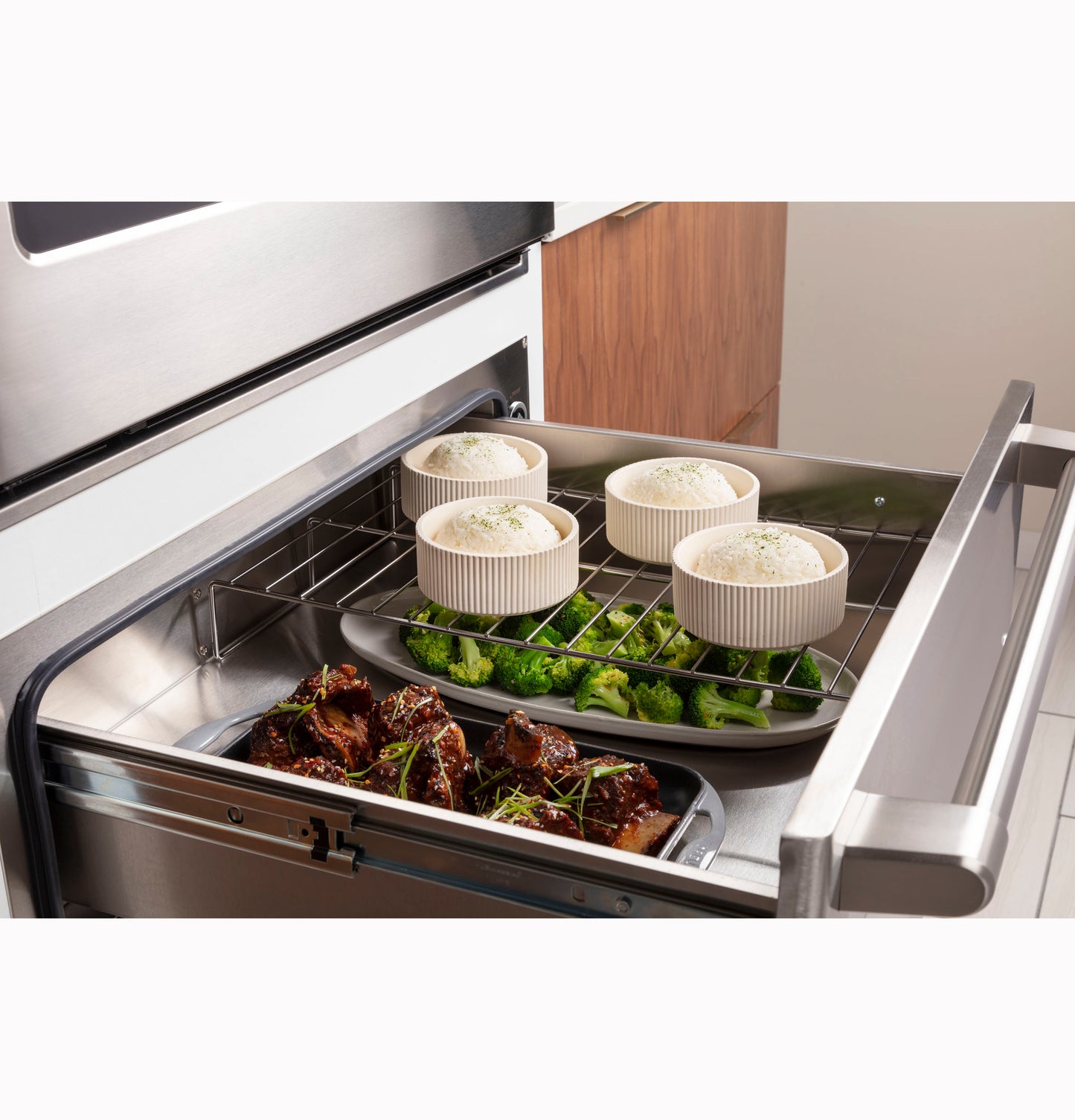 Metal warming drawer with ribs and broccoli on plate and four ramekins of dough in kitchen and wall ovens setting
