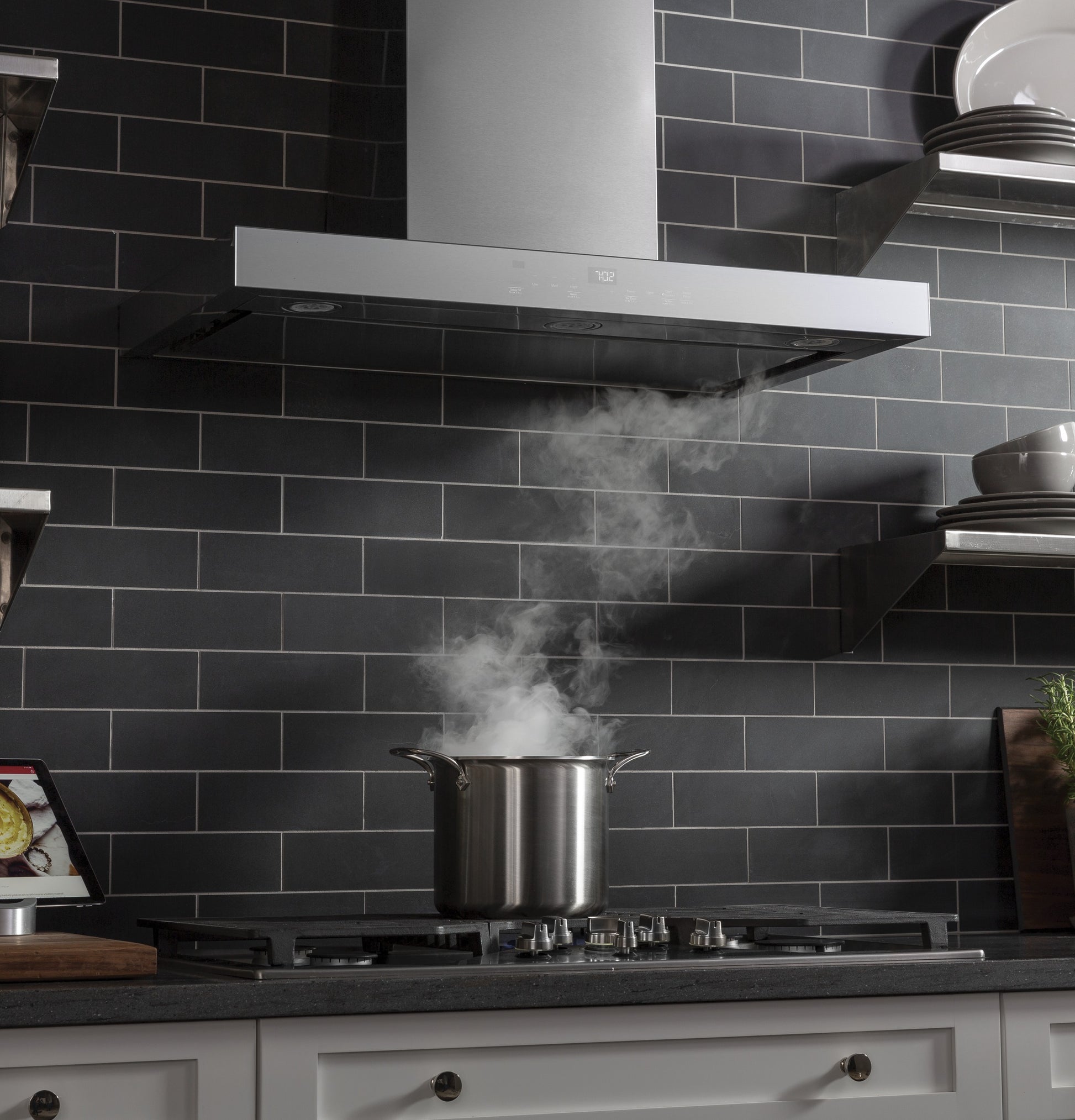 Modern kitchen with stainless steel wall mount vent hood extracting steam from pot on gas cooktop with dark tile backsplash