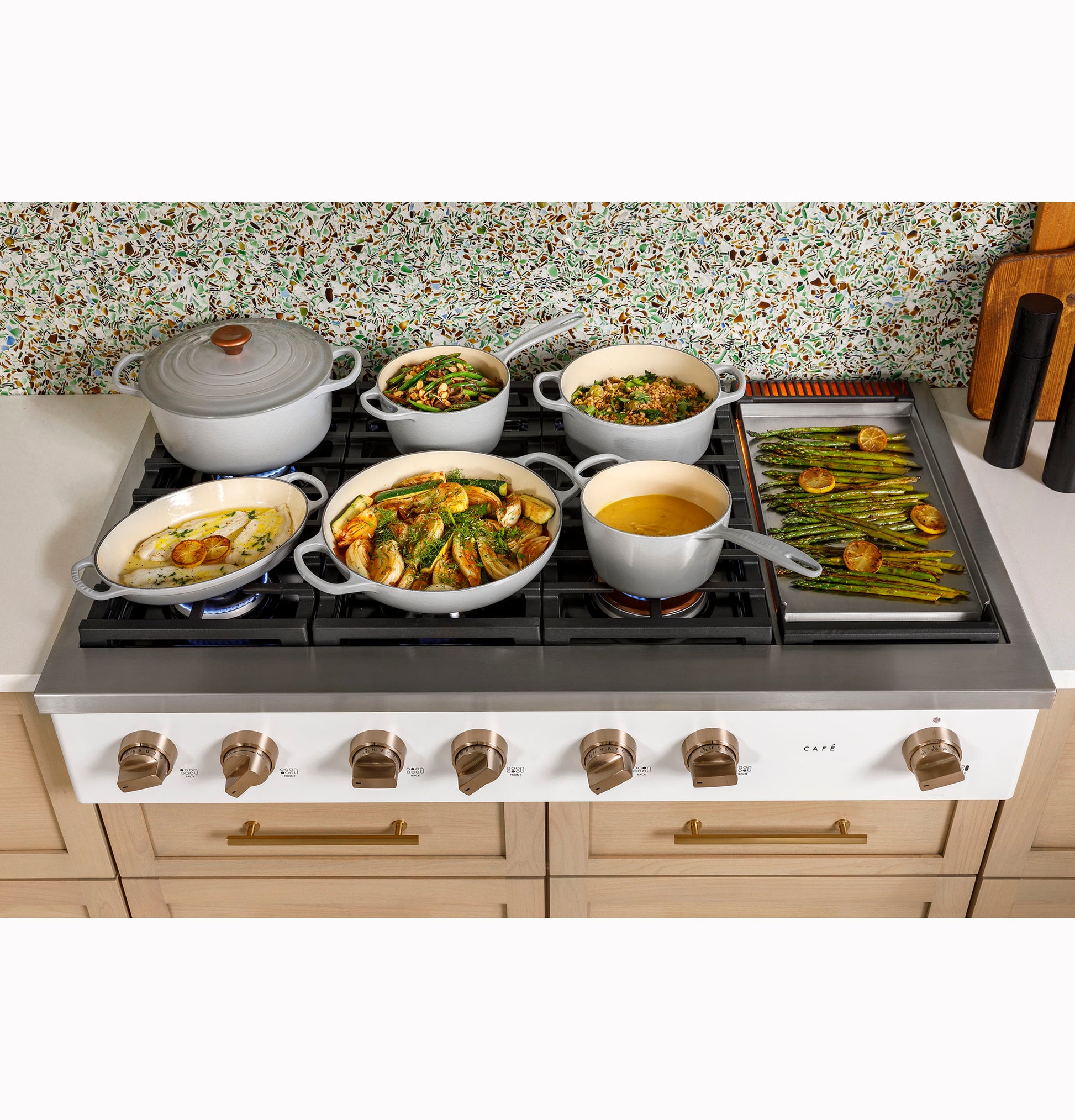 Commercial-style gas rangetop with six burners and integrated griddle cooking various vegetables and soups in kitchen setting