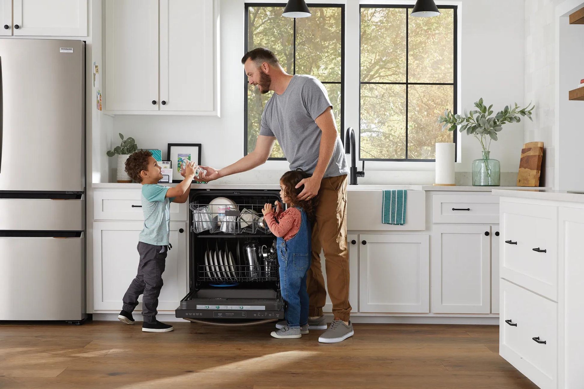 Man unloading open built-in dishwasher with two children in bright modern kitchen with stainless appliances