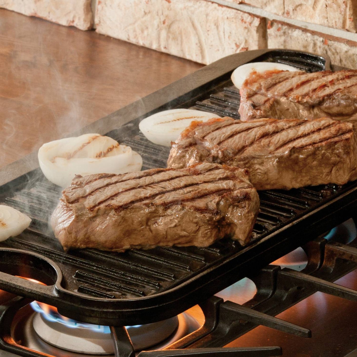Cast iron griddle grilling seasoned steaks and sliced onions over gas stovetop burners in cookware and outdoor living collections
