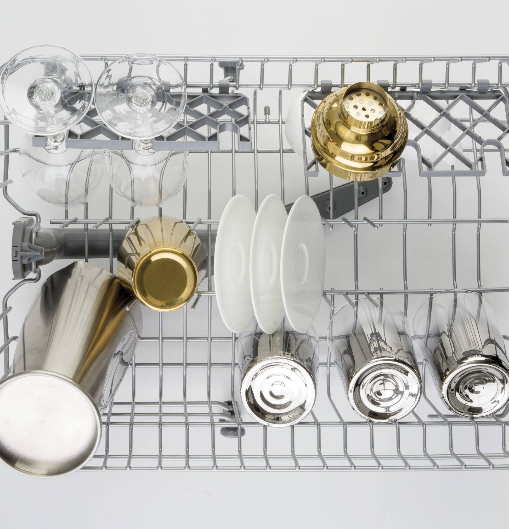 Top view of wine glasses, cocktail shaker, white plates, and bar tools arranged in dishwasher rack for kitchenware cleaning