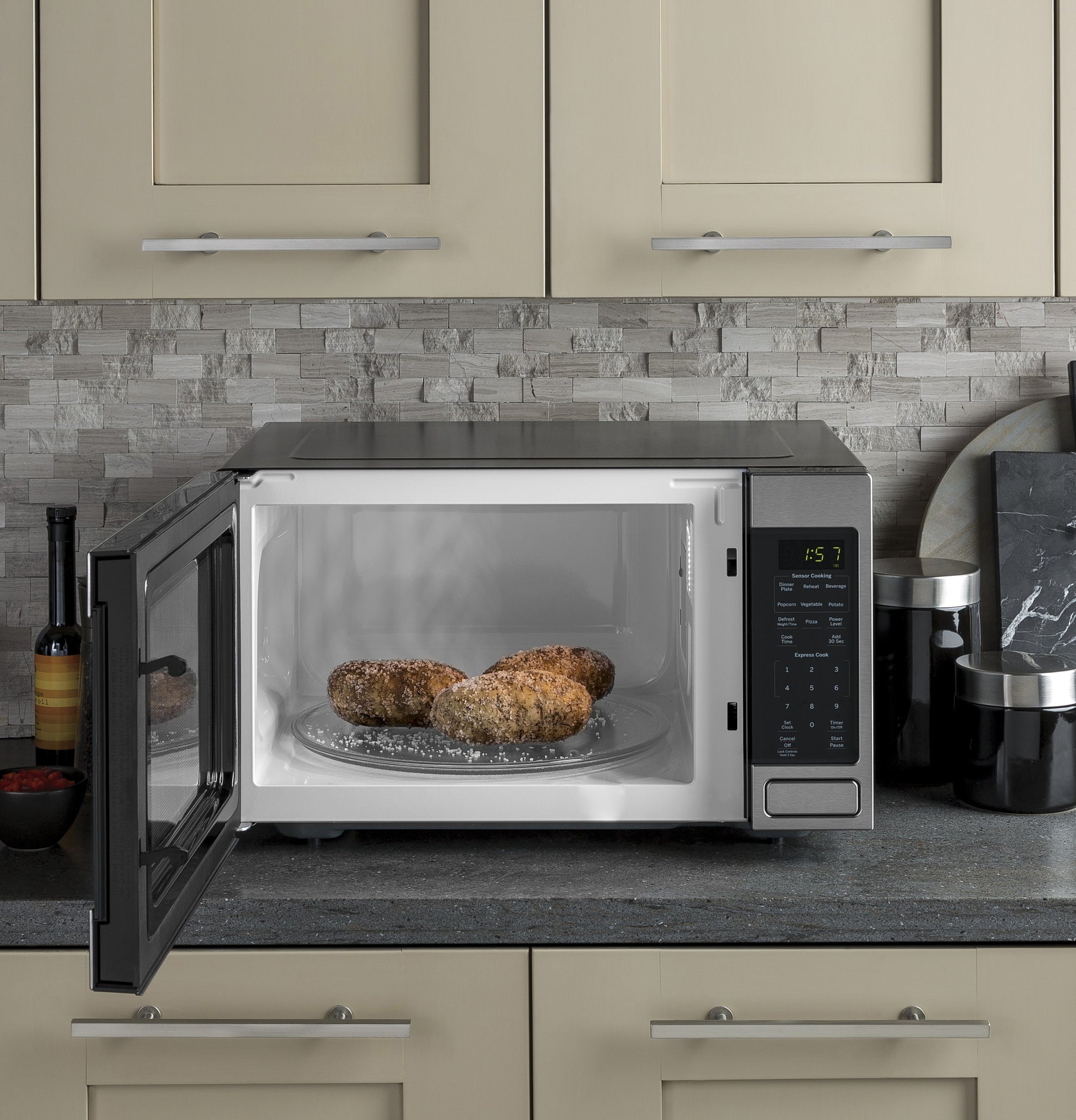 Countertop microwave with open door heating three baked potatoes on rotating glass tray in modern kitchen setting
