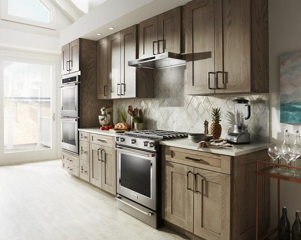 Stainless steel low profile under-cabinet ventilation hood installed above a stainless gas range in a kitchen with light wood cabinets, part of Under Cabinet Range Hoods and Kitchen & Kitchenware collections.