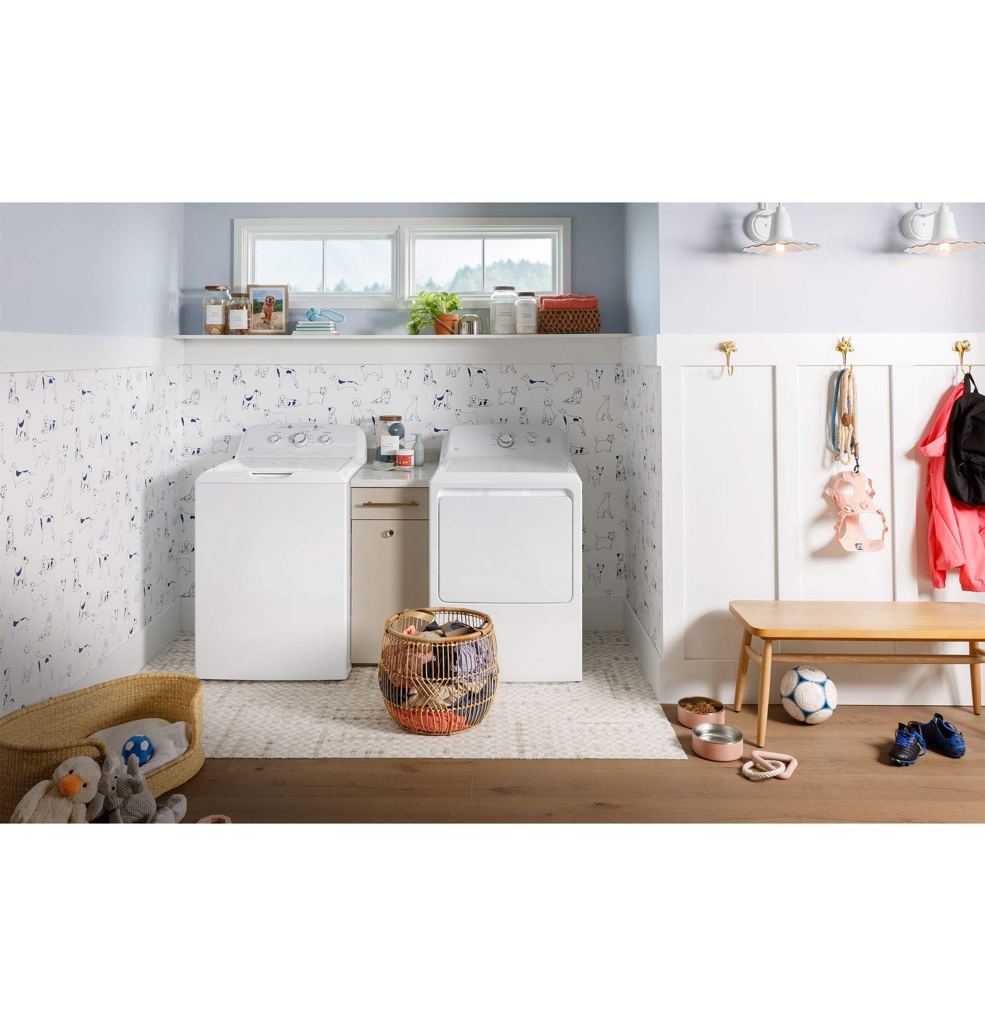 Laundry room scene with white washer and electric dryer set, pet bed, toys, and bench with hanging coats and bags