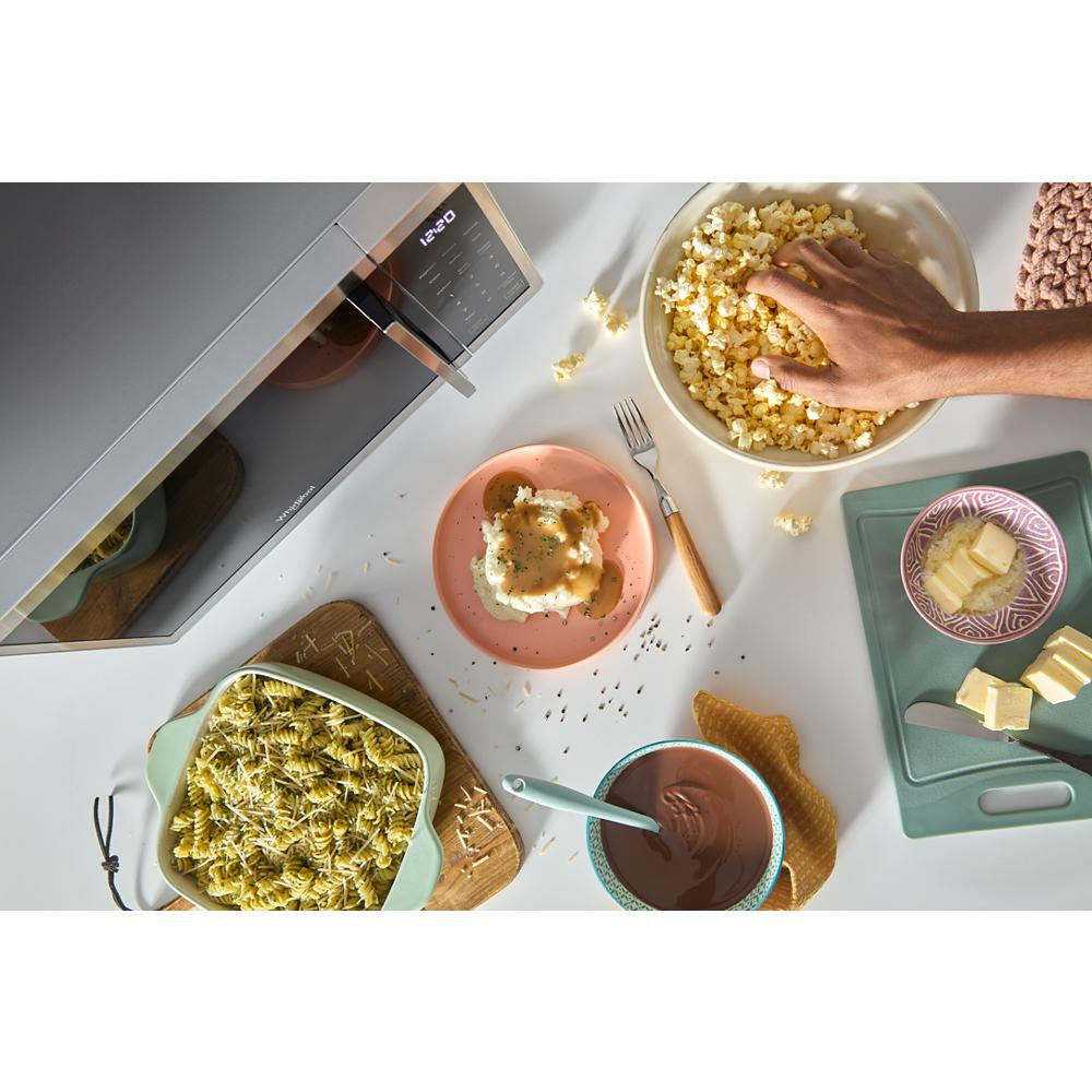 Black countertop microwave open on white kitchen counter with bowls of popcorn, pasta, and melted chocolate nearby