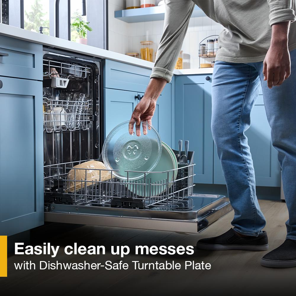 Person placing glass microwave turntable plate into open dishwasher in kitchen with blue cabinets and natural light