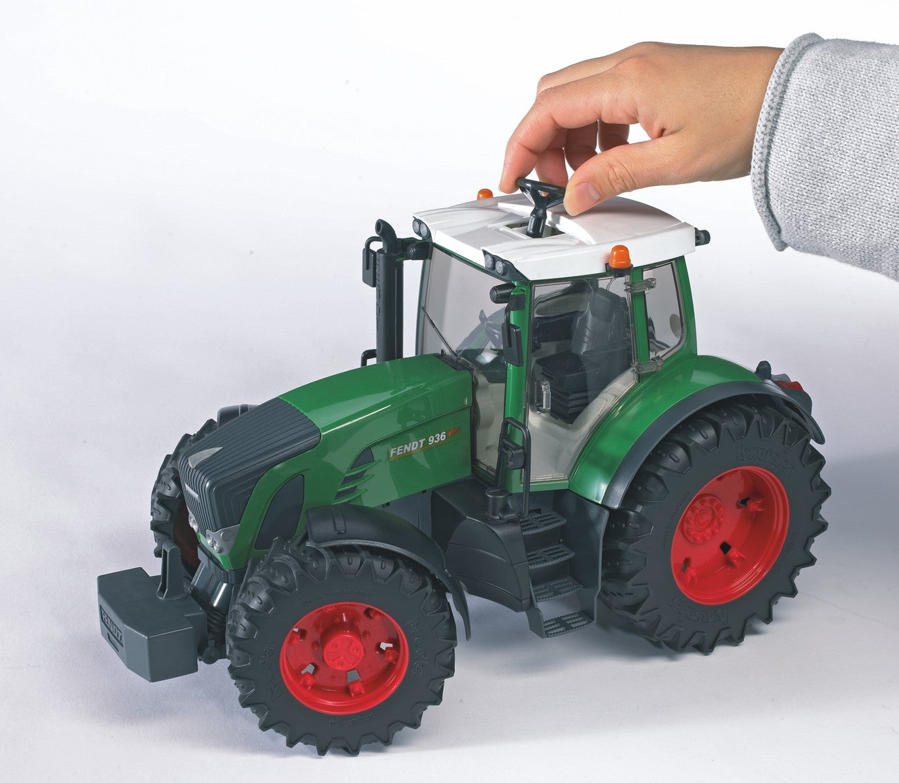 Green toy tractor with red wheels and detailed cabin, being adjusted by a hand wearing a gray sleeve