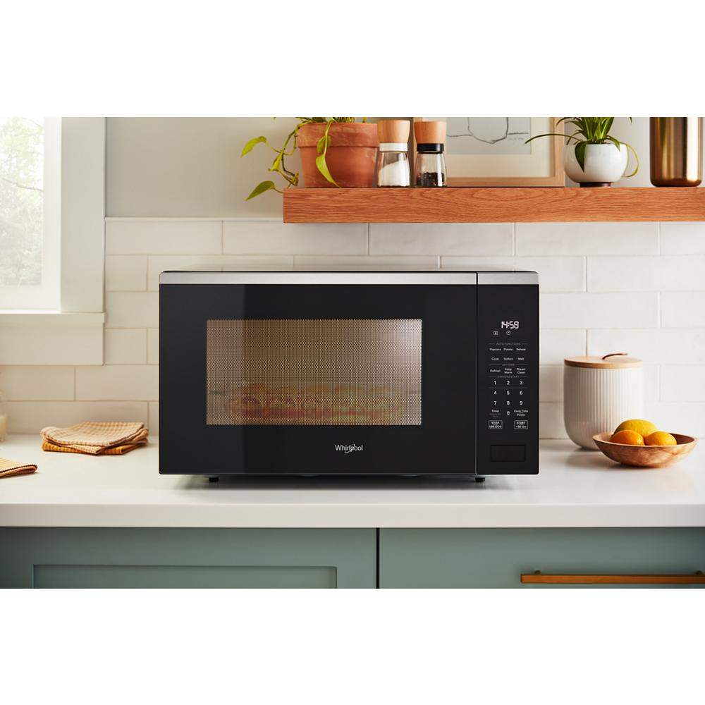 Black sensor microwave with digital control panel on white kitchen countertop near green cabinetry and wood shelf decor