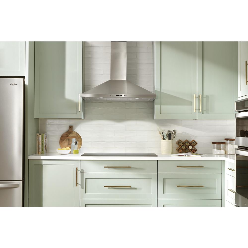 Stainless steel chimney hood above cooktop in kitchen with sage green cabinetry and built-in double oven appliances