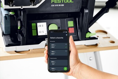 Hand holding smartphone displaying settings while adjusting a Festool cordless table saw in a bright workshop