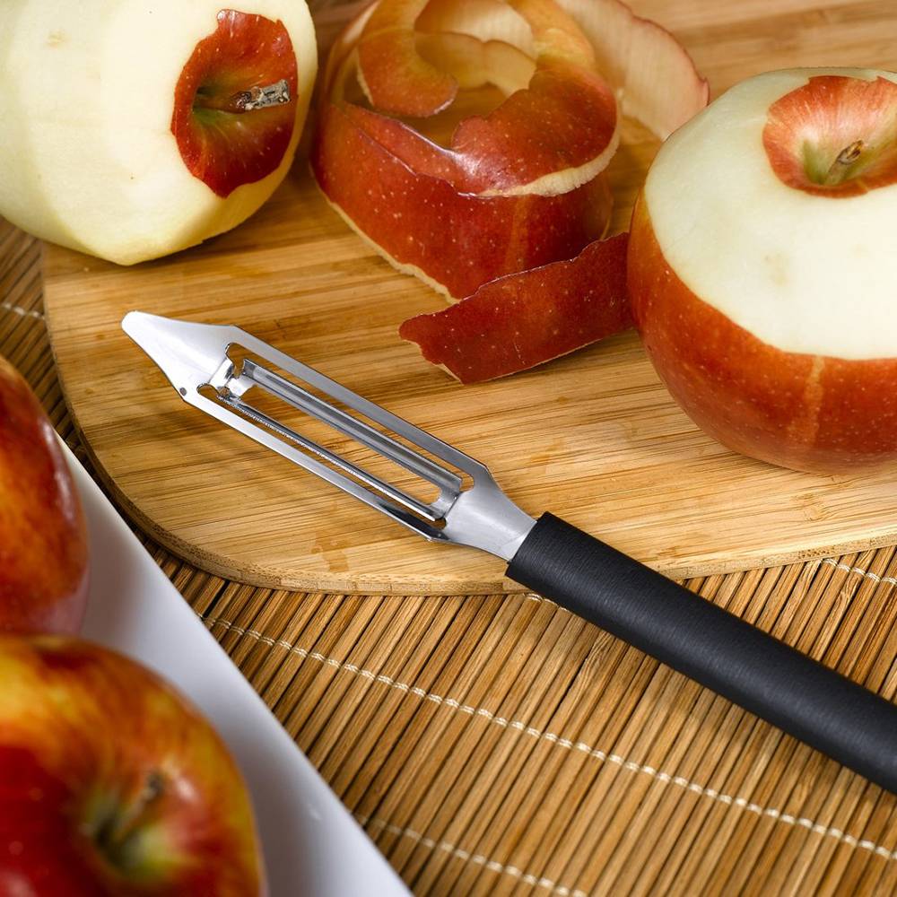 Stainless steel vegetable peeler with black resin handle next to peeled apples and apple skin on cutting board in Kitchenware collection