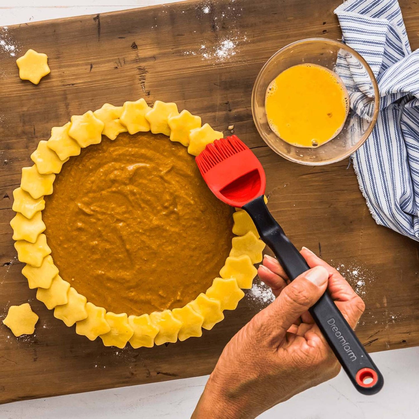 Hand holding red silicone basting brush with black handle applying egg wash to star-shaped dough pieces on wooden surface, Kitchenware