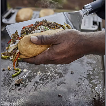 Hand holding stainless steel griddle spatula loading cooked sandwich filling on toasted bread for outdoor grilling tools use