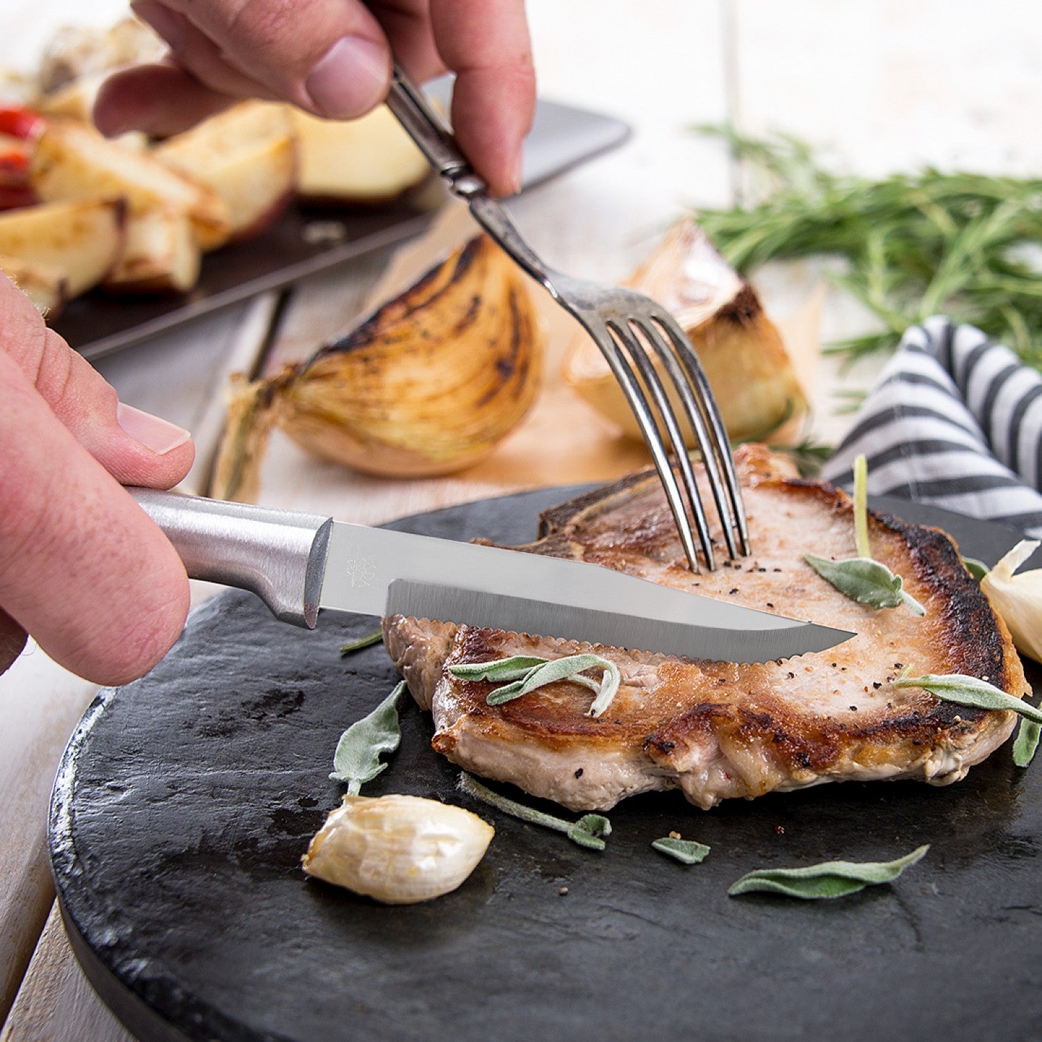 Serrated steak knife with solid aluminum handle slicing grilled pork chop on a black plate with garlic and fresh herbs, ideal for Kitchenware and Cooking Utensils under $50.