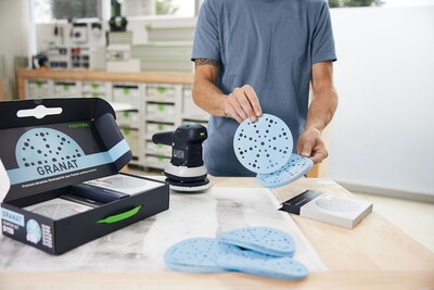 Person handling blue sanding discs from Festool Granat D150 starter set with sander on workbench for home improvement tasks
