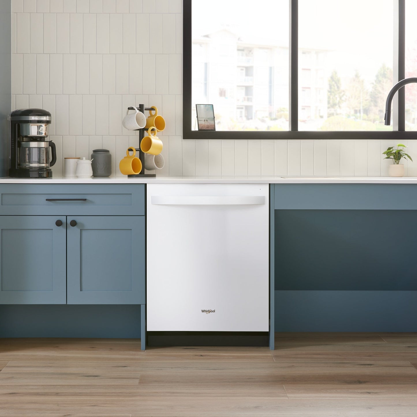 White Whirlpool dishwasher integrated into blue lower kitchen cabinets with a coffee maker and mug rack nearby