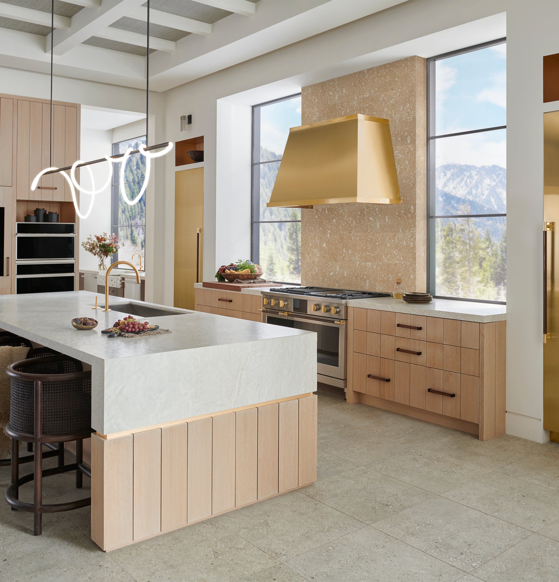 Modern kitchen featuring a brass designer range hood above stove, with light wood cabinetry and large windows in kitchenware collection