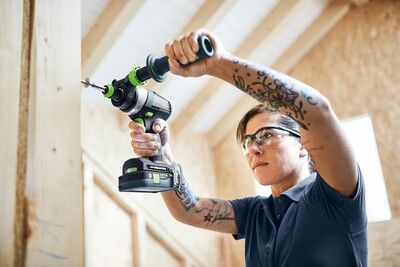Person wearing safety glasses using a cordless hammer drill to insert a screw into wooden construction framing