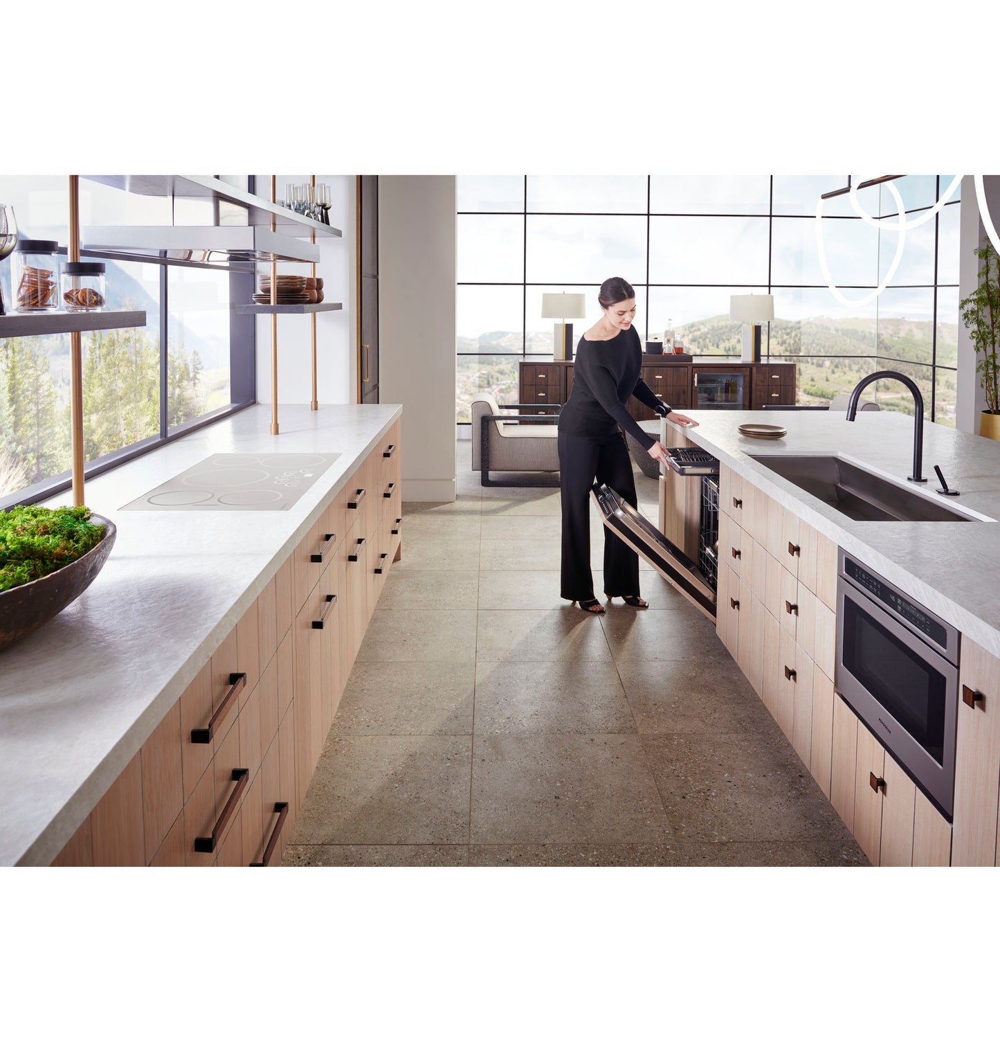 Woman opening stainless steel microwave drawer installed in light wood kitchen cabinetry with large windows and island sink