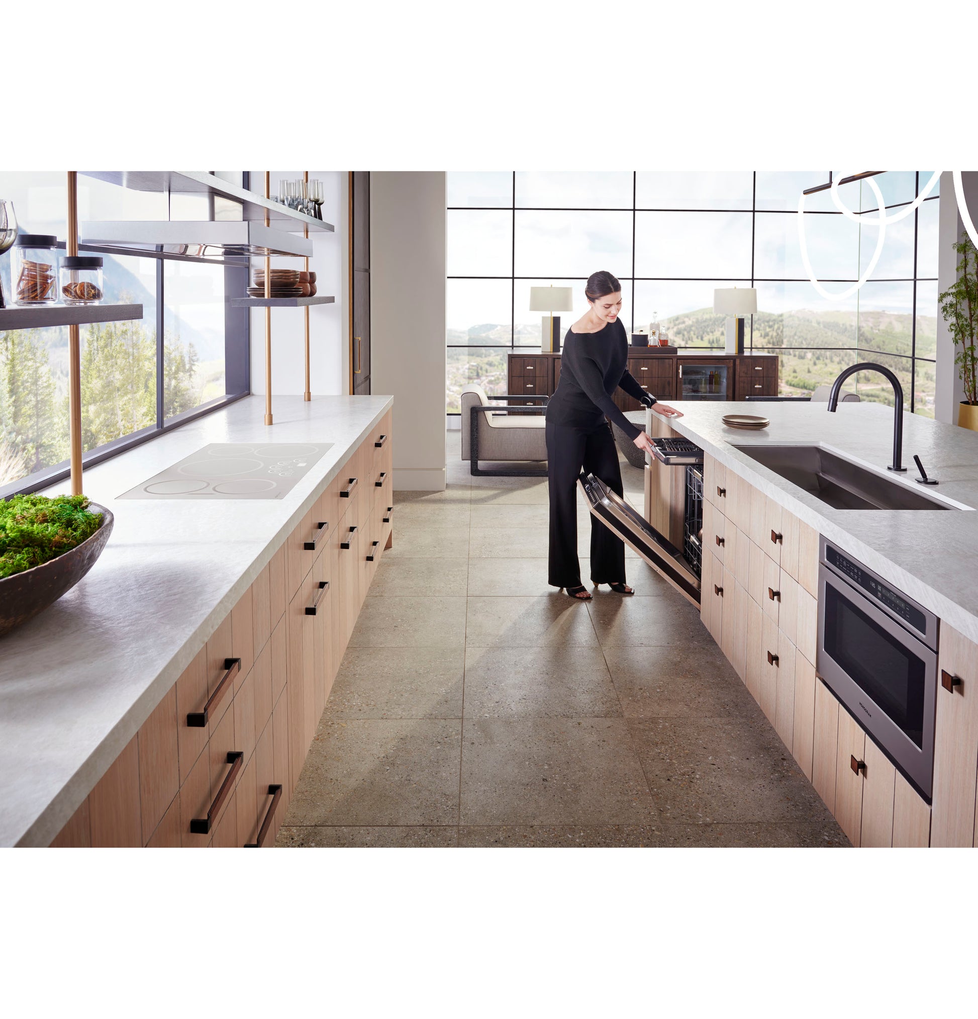 Woman opening stainless steel microwave drawer installed in light wood kitchen cabinetry with large windows and island sink