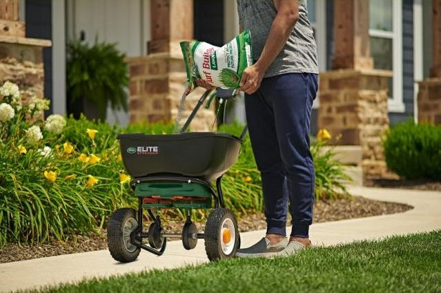 Person pouring lawn fertilizer into a black wheeled spreader on a residential garden path
