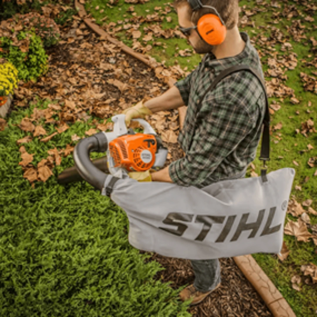 Person wearing ear protection using an orange gas leaf blower with attached collection bag in a garden setting
