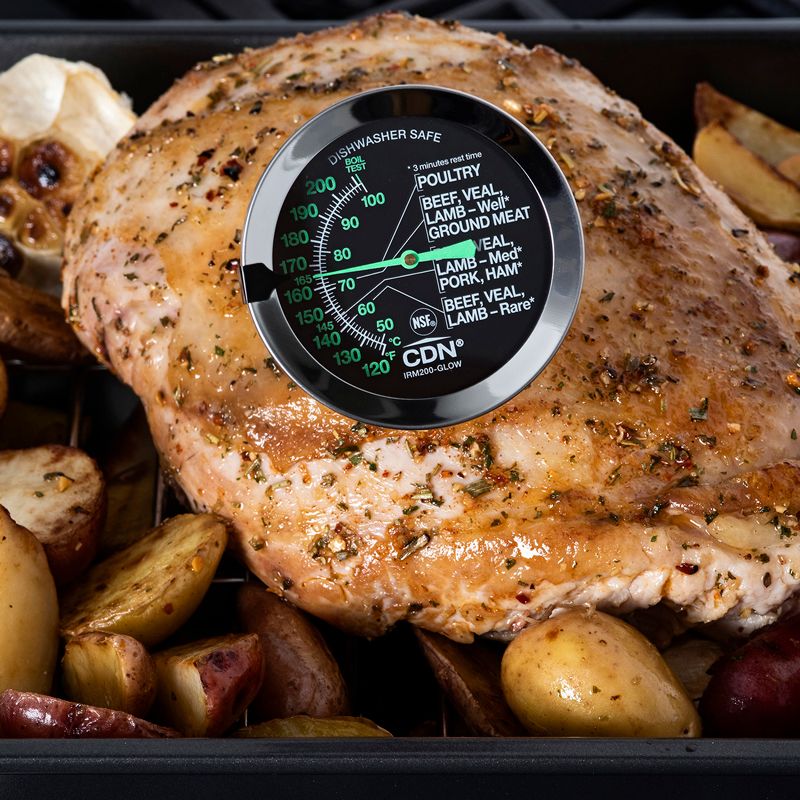 Ovenproof glow dial thermometer inserted in roast chicken surrounded by roasted potatoes in kitchenware setting