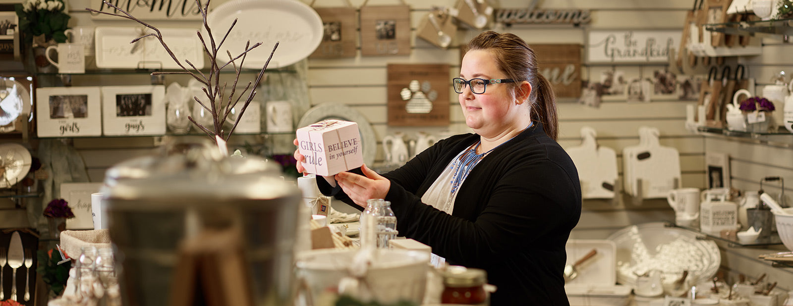 Woman holding a decorative wooden block with inspirational text inside a cozy shop filled with rustic home decor and gifts.