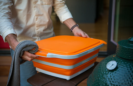 Person holding collapsible orange BBQ prep tub with lid on outdoor table near green ceramic grill