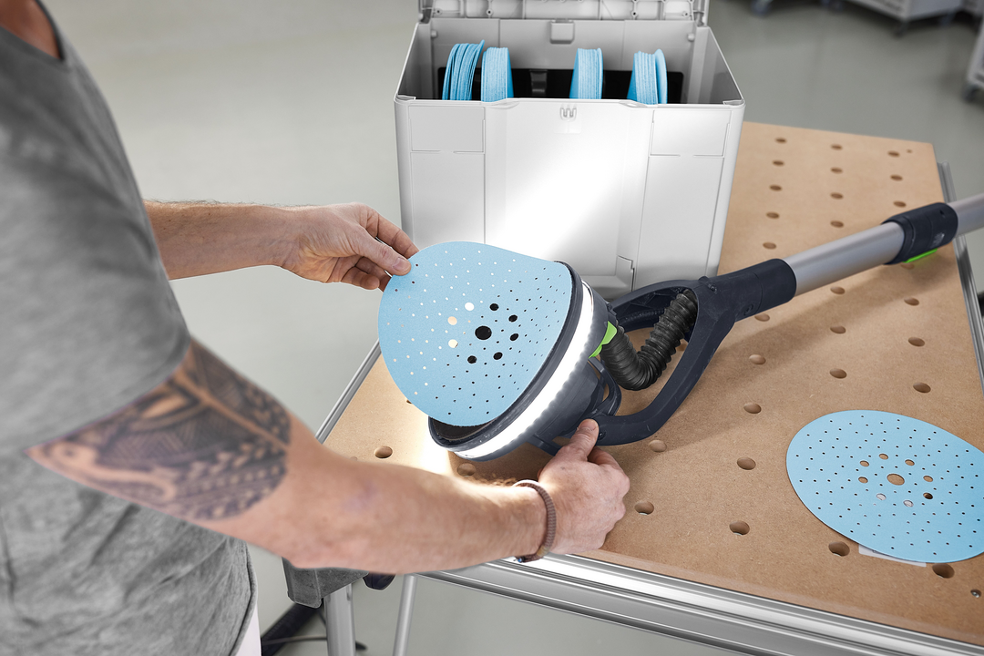 Man attaching blue sanding disc to long-reach sander near white Festool Granat abrasives storage box in workshop