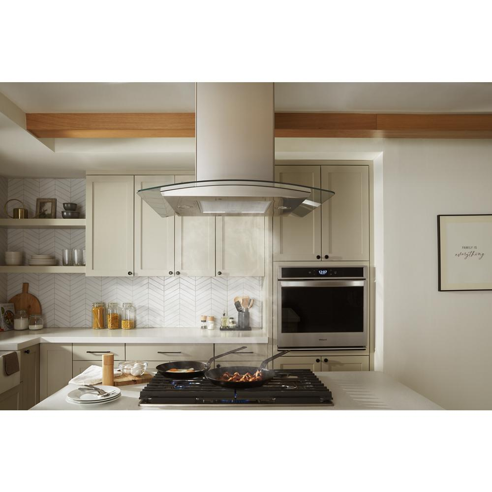 Modern kitchen with black cooktop featuring two pans cooking over flames beneath a curved glass range hood and white cabinetry