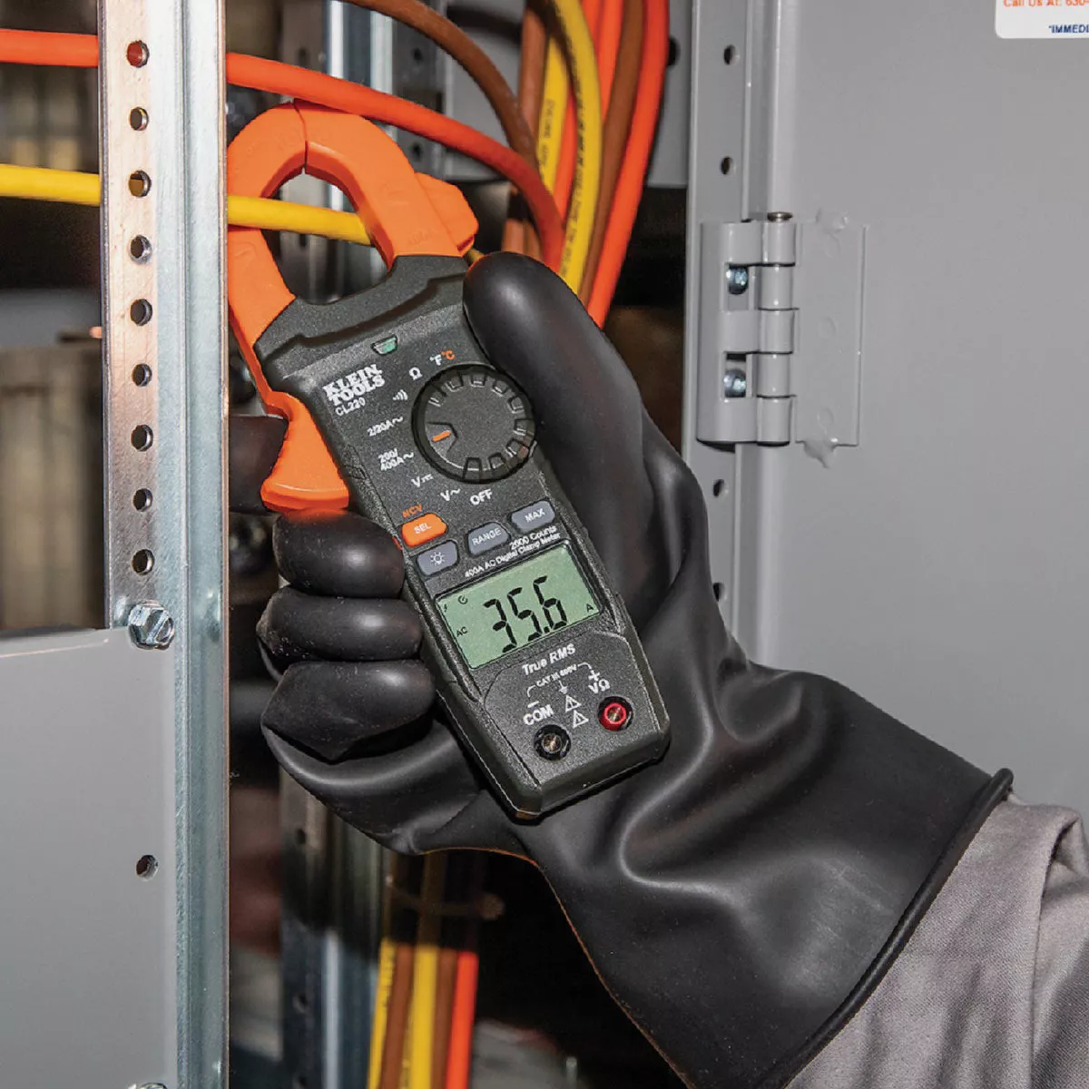 Hand in protective glove holding clamp meter measuring current on orange insulated electrical wire in panel box