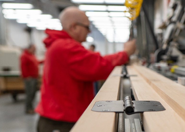 Close-up of a metal door hinge attached to wooden boards with a blurred background showing people working in a workshop or home improvement environment.