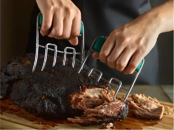 Hands using two meat shredding claws with green handles to pull apart a large cooked piece of meat on a wooden cutting board.