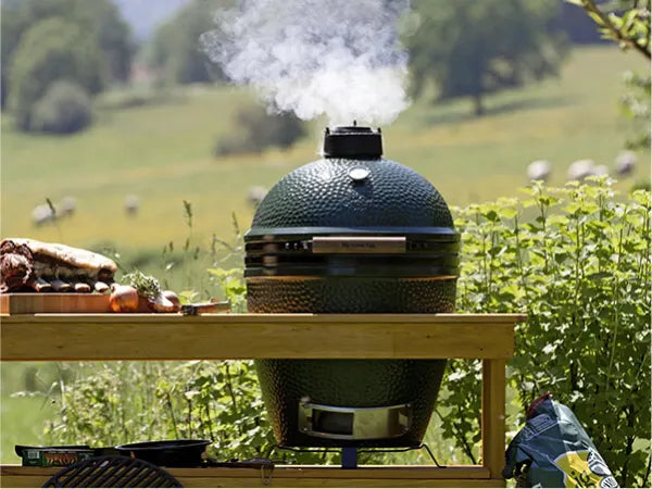 Green ceramic outdoor grill with smoke rising from the vent, placed on a wooden table with cooked meat and fresh vegetables nearby in a garden setting.