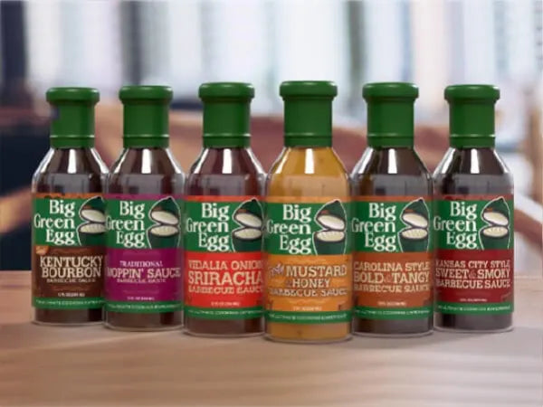 Six assorted barbecue sauce bottles with green caps and branded Big Green Egg labels, arranged in a row on a wooden surface.