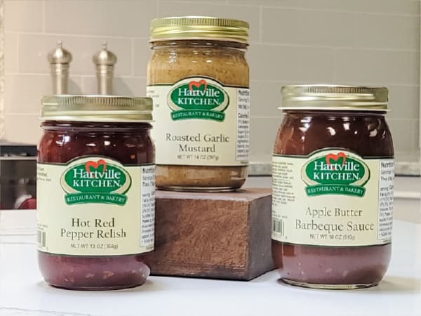 Three jars of Hartville Kitchen condiments including hot red pepper relish, roasted garlic mustard, and apple butter barbeque sauce displayed on a kitchen counter.