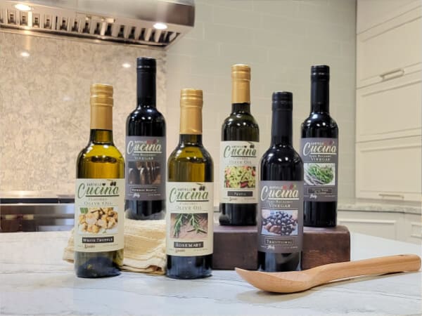 Six glass bottles of flavored oils and vinegars labeled with different culinary herbs, arranged on a kitchen countertop near a wooden spoon.