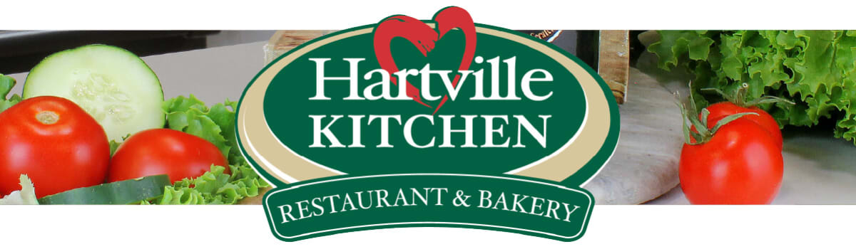 Green and beige Hartville Kitchen logo featuring a red heart and banner reading Restaurant & Bakery, set against fresh vegetables including tomatoes, cucumber, and lettuce.