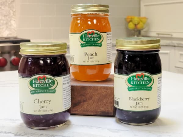 Three jars of Hartville Kitchen jams including cherry, peach, and blackberry flavors displayed on a kitchen countertop with white cabinetry and fruit bowl in the background.