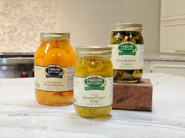 Three jars of preserved foods including peeled peach halves, banana pepper rings, and bread and butter pickles displayed on a kitchen counter with white cabinetry in the background.