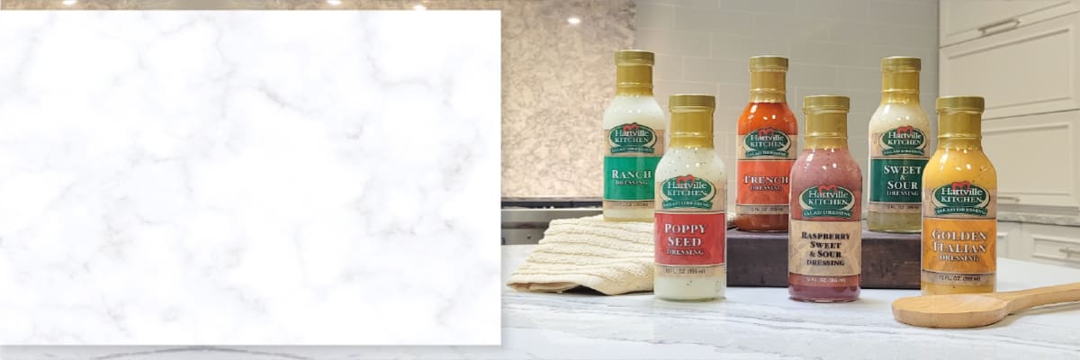 Five glass bottles of salad dressing in various flavors including poppy seed and raspberry vinaigrette arranged on a kitchen counter with white cabinetry in the background.