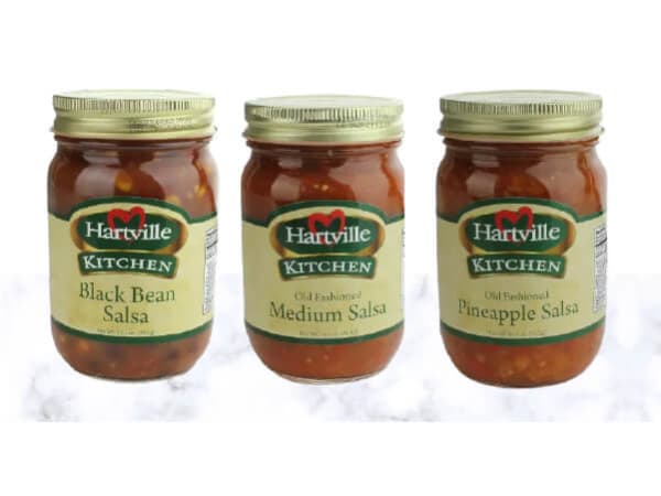 Three glass jars of Hartville Kitchen salsa varieties including black bean, medium, and pineapple flavors, arranged side by side on a white surface.