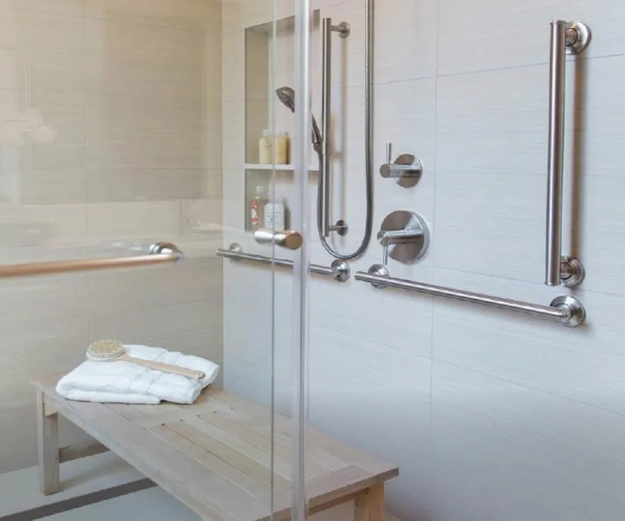 Shower area with light tile walls featuring multiple horizontal and vertical stainless steel safety grab bars, a wooden bench holding folded towels and bath brushes.
