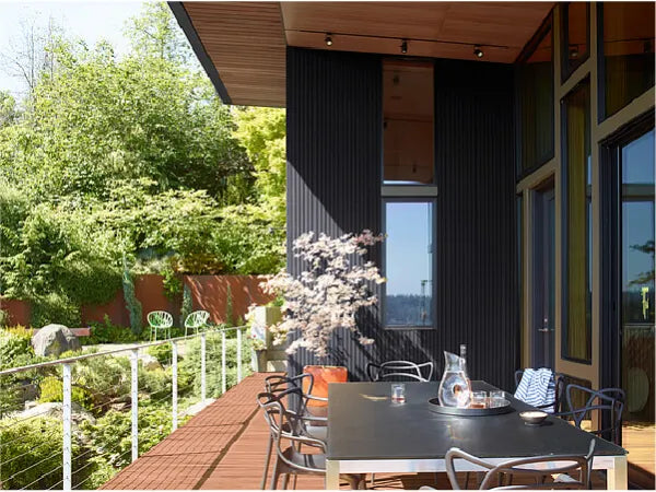Outdoor dining area with a long table, modern chairs, glass railing, and lush green trees surrounding a wooden deck on a sunny day.